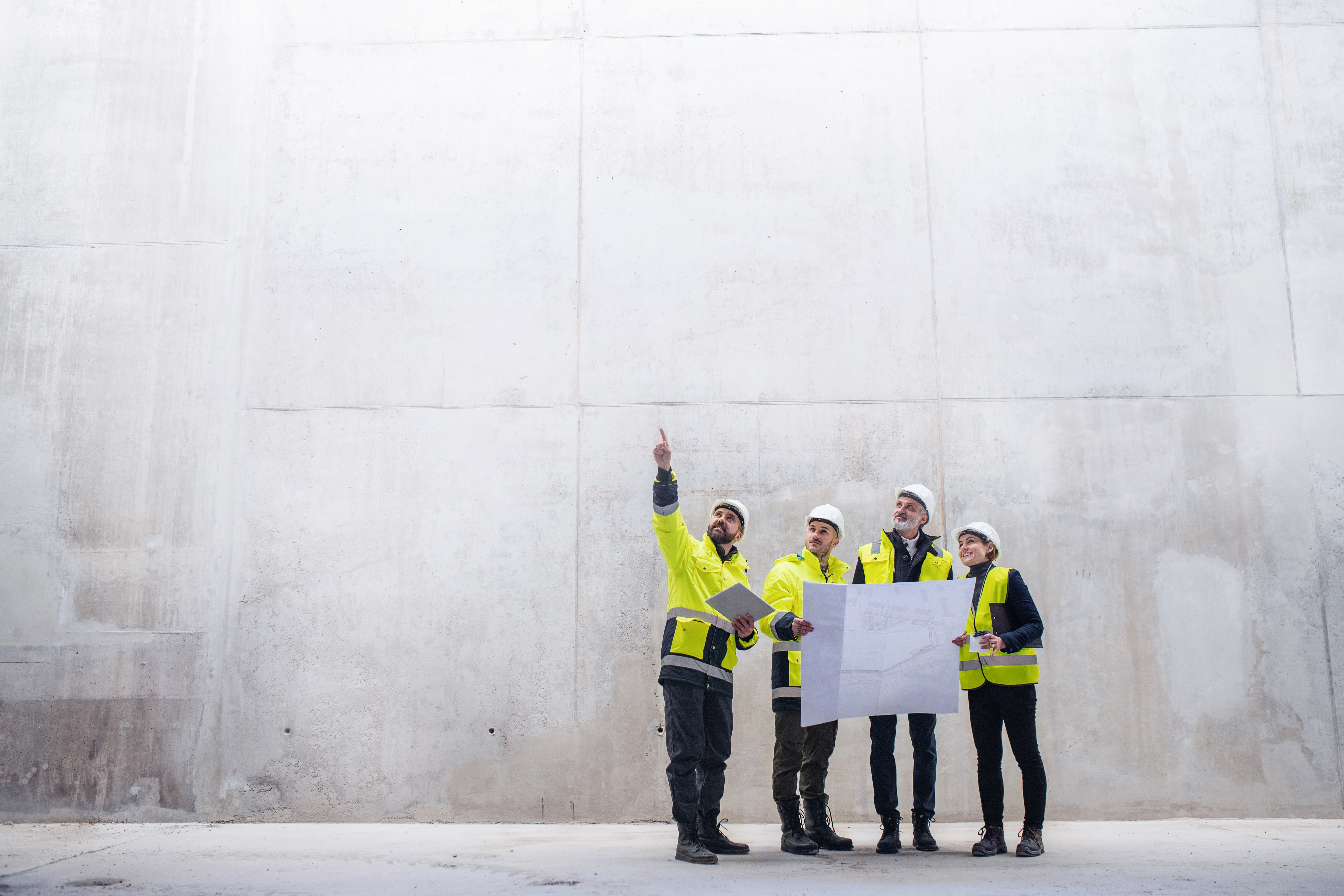Vier Menschen in Warnkleidung und Schutzhelmen stehen mit einem Bauplan in der Hand vor einer hohen Betonwand.