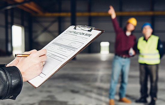 Eine Person füllt eine Gefährdungsbeurteilung aus, während ein Bauherr einer Aufsichtsperson die Baustelle zeigt