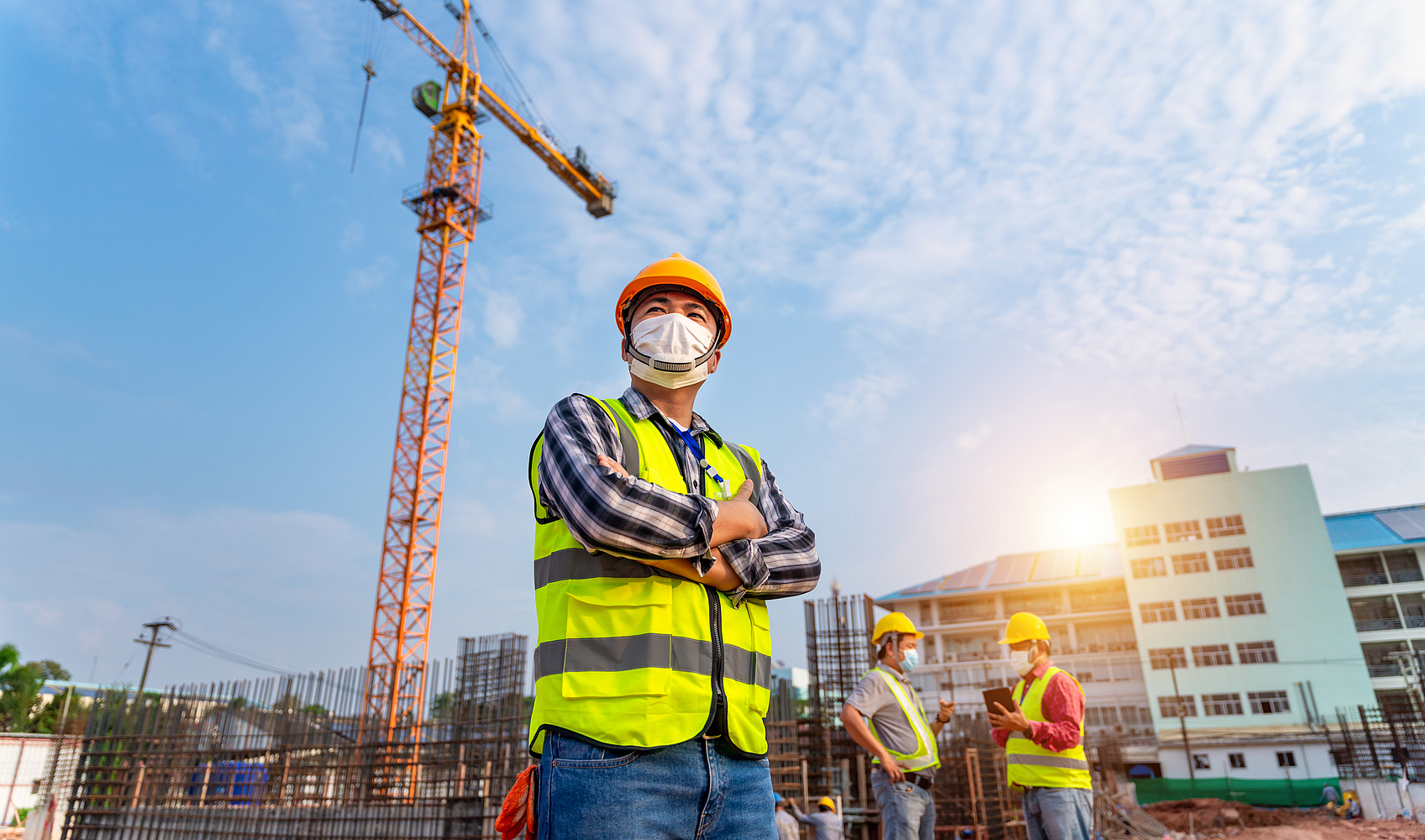 Drei Männer mit Schutzhelmen und Mund-Nase-Bedeckung stehen auf einer Baustelle. Im Hintergrund steht ein Kran.